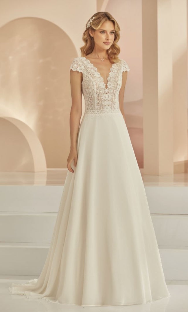 Eine elegante Braut im „Clara“ A-Linien-Brautkleid von „Susannas Brautmode“, verziert mit feiner Spitze und einem fließenden Rock, posiert vor einem ruhigen Hintergrund und strahlt klassische Schönheit aus.