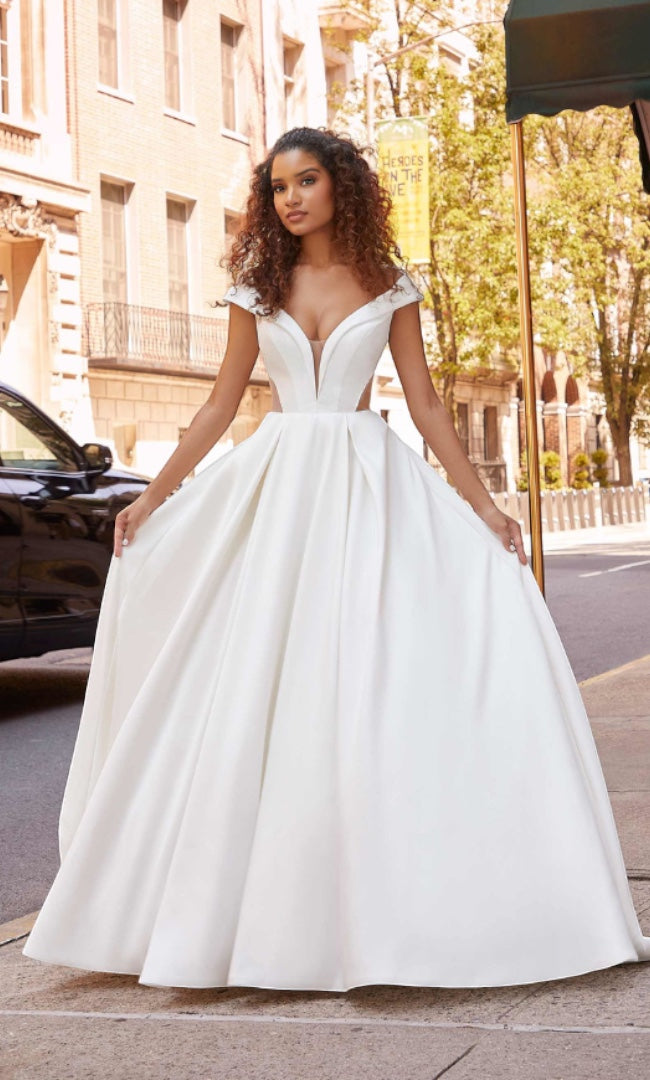 Eine Braut in einem schicken „Isabella“ A-Linien-Brautkleid von „Susannas Brautmode“ mit tiefem V-Ausschnitt, die auf einer städtischen Straße steht und ein Bild moderner Eleganz darstellt.