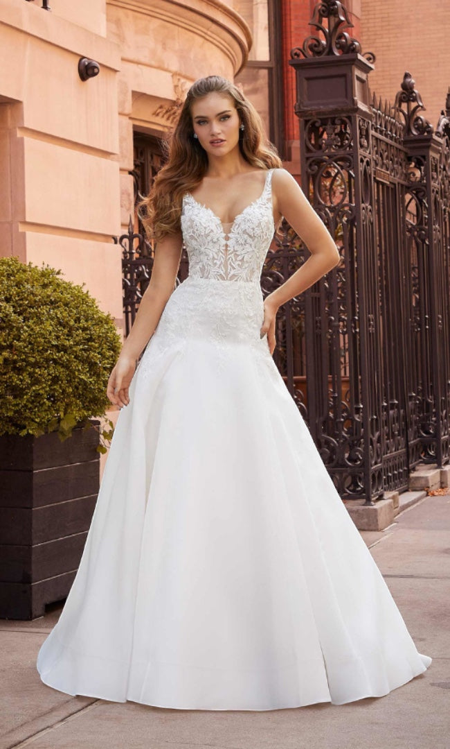 Eine strahlende Braut  trägt das „Angelina“ A-Linien-Brautkleid von „Susannas Brautmode“, mit einem tiefen V-Ausschnitt und aufwendigen Spitzen, steht stolz auf einer urbanen Straße und strahlt zeitlose Eleganz aus.