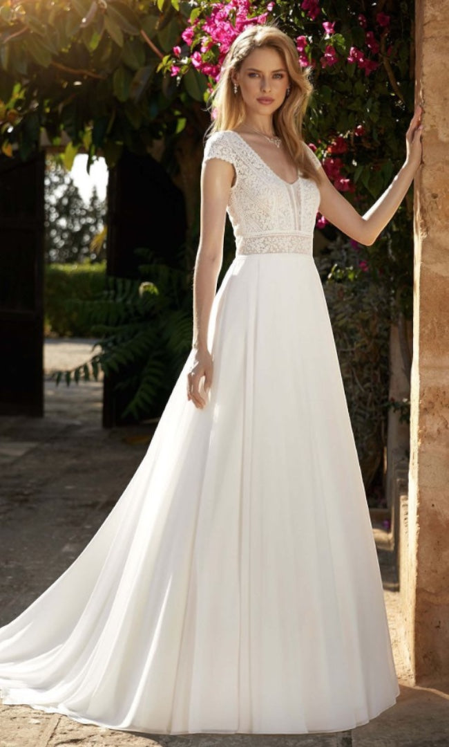 Eine Braut in einem schlichten und eleganten A-Linien Hochzeitskleid mit zarten Spitzenärmeln und einem V-Ausschnitt, stehend in einer sonnigen Allee.