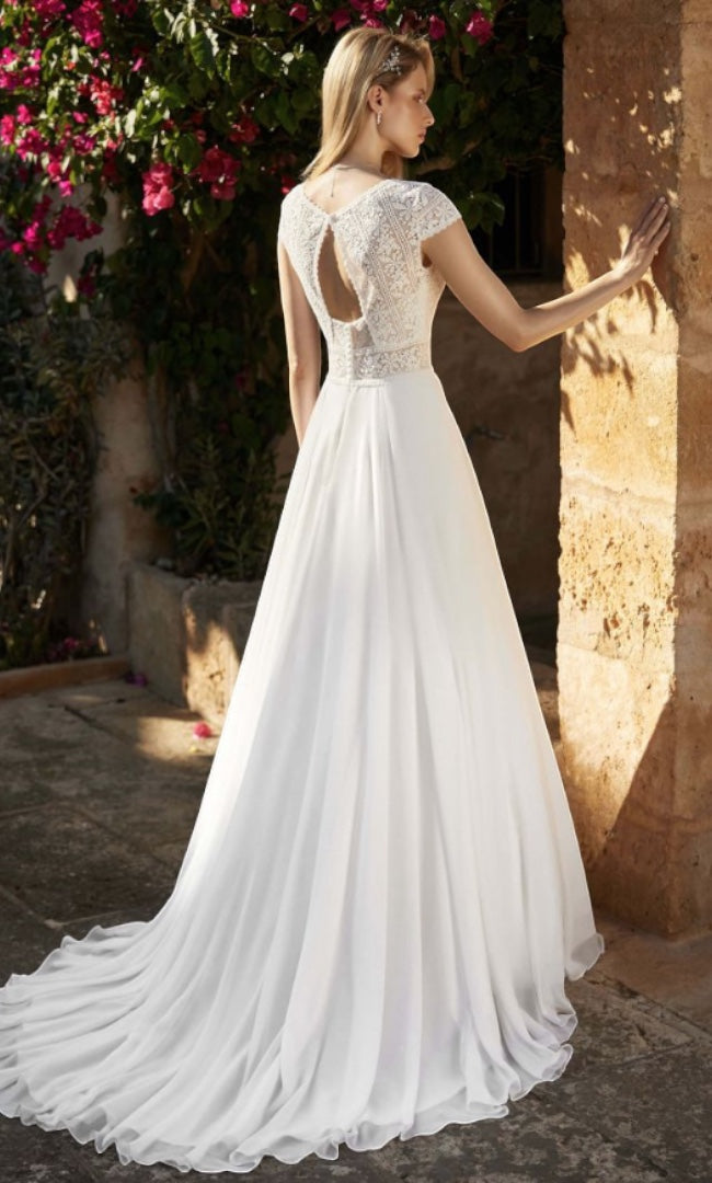 Eine Braut in einem A-Linien Hochzeitskleid mit filigranem Spitzenrücken und einem eleganten, weiten Rock mit einer kleinen Schleppe, umgeben von blühenden Büschen.